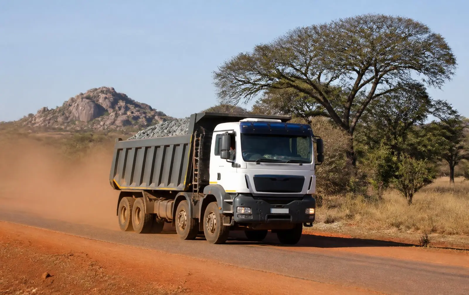 Loaded tipper truck hauling bulk minerals along a red-earth highway past granite kopjes in Zimbabwe